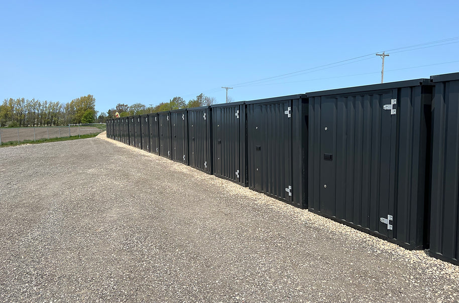  Flatpack containers in black colour  with secure locks, positioned side-by-side on a gravel surface under a clear blue sky.